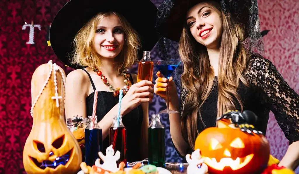 Two women in witch costumes celebrating with Halloween drinks for adults, surrounded by spooky decorations.
